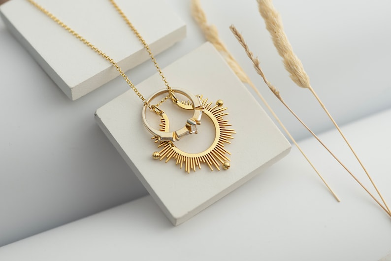 May include: A gold necklace featuring a sunburst pendant with two rings attached. The rings are made of gold and silver.