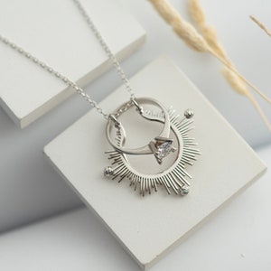 May include: A silver necklace with a pendant featuring two interlocking rings and a single diamond. The pendant is surrounded by a sunburst design.