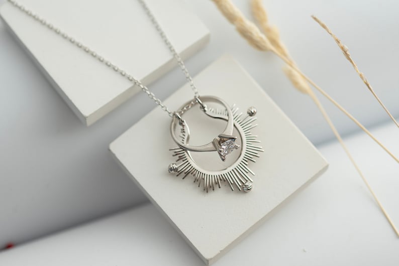 May include: A silver necklace with a pendant featuring two interlocking rings and a single diamond. The pendant is surrounded by a sunburst design.