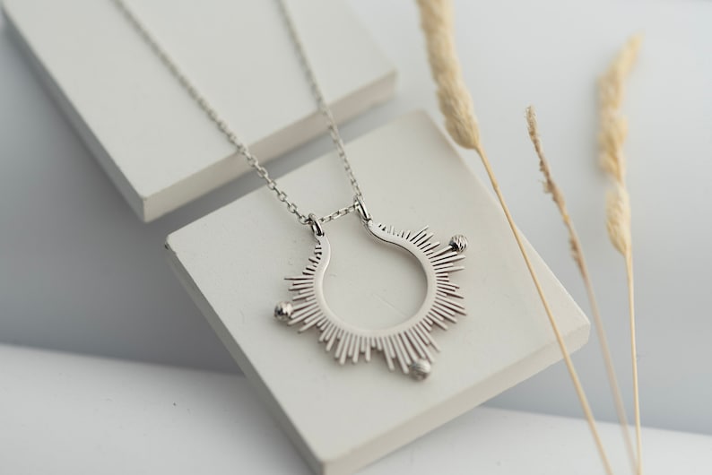 May include: A silver necklace featuring a sunburst pendant. The pendant is a circle with rays radiating outwards, each ending in a small bead.