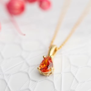 May include: A gold pendant necklace featuring a pear-shaped, orange gemstone. The gemstone is set in a gold bezel and hangs from a delicate gold chain. The necklace is displayed on a white, textured surface.