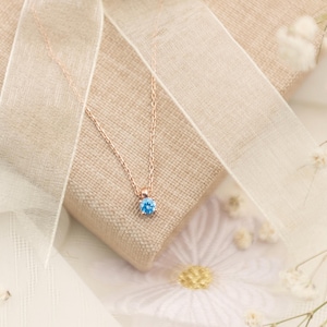 Könnte beinhalten: Eine zarte Halskette aus Roségold mit einem runden, hellblauen Edelsteinanhänger. Die Halskette liegt auf einem beigen Stoff, akzentuiert durch ein transparentes Band und kleine weiße Blüten, was eine weiche, elegante Ästhetik erzeugt.