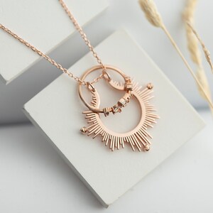 May include: A rose gold necklace featuring a sunburst pendant and a smaller circle pendant with small clear stones. The necklace is displayed on a white surface.