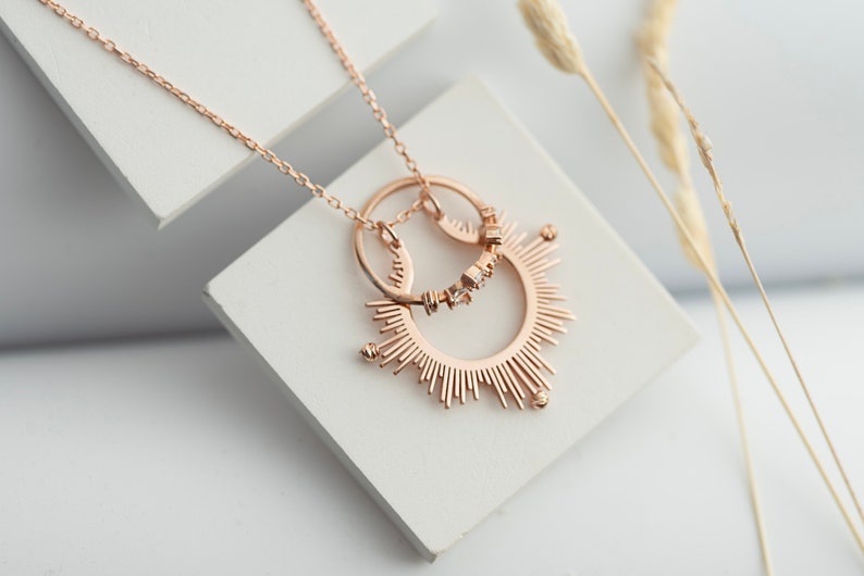 May include: A rose gold necklace featuring a sunburst pendant and a smaller circle pendant with small clear stones. The necklace is displayed on a white surface.