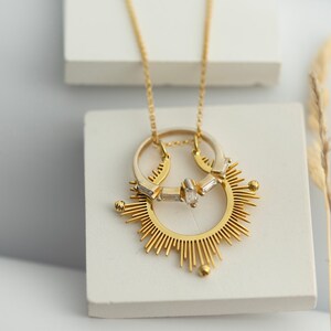 May include: A gold necklace with a sunburst pendant featuring two interlocking rings with a single diamond. The necklace is displayed on a white surface.