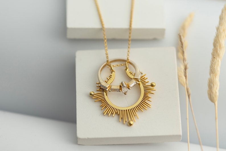 May include: A gold necklace with a sunburst pendant featuring two interlocking rings with a single diamond. The necklace is displayed on a white surface.