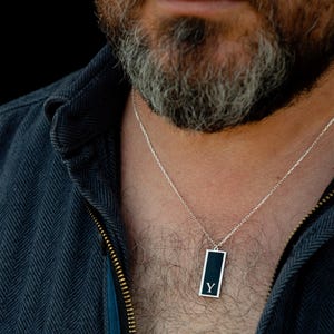 Può includere: Collana d'argento con un ciondolo rettangolare. Il ciondolo ha uno sfondo nero con la lettera "Y" in bianco. La collana è indossata sopra una camicia blu scuro.