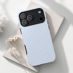 May include: A light blue phone case with a repeating pattern of small red dots. The case has a black camera module and black trim. The phone case is on a white surface.