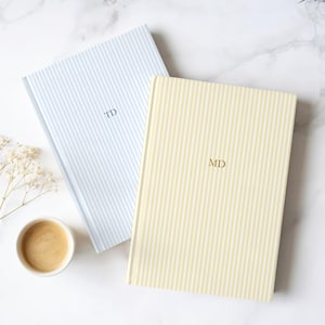 Può includere: Due quaderni con copertina rigida a righe verticali. Uno è azzurro chiaro con le iniziali "TD" e l'altro è giallo con le iniziali "MD". Sono presenti anche una tazza di caffè e rametti di fiori bianchi.