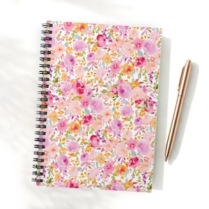 Può includere: Un quaderno a spirale con un motivo floreale nei toni del rosa, pesca e giallo. Una penna color oro rosa è appoggiata accanto al quaderno su una superficie bianca. La copertina presenta un design ad acquerello di fiori e foglie.