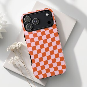 May include: A phone case featuring a bright orange and pale pink chequered design. The case has a black camera section and a glossy finish. The pattern is a striking, attention-grabbing feature.
