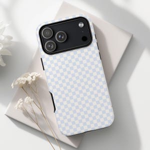 May include: A smartphone case featuring a light blue and white chequered design. The case has a black camera module and black edges. The case is on a white surface with dried flowers.