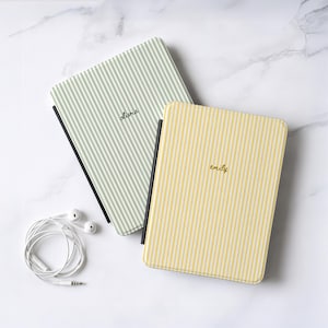 May include: Two rectangular e-reader cases with vertical pinstripes, one in sage green with the name olivia and one in yellow with the name emily, accompanied by a pair of white wired earbuds with a standard 3.5mm jack connector.