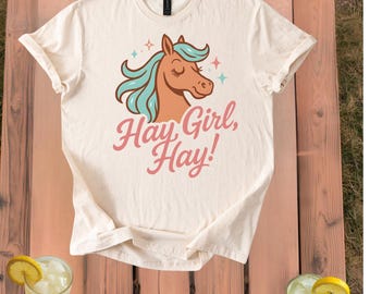 May include: Cream-colored t-shirt featuring a cartoon horse with teal hair and the text "Hay Girl, Hay!" in pink. The horse has closed eyes and is surrounded by stars. The shirt is displayed on a wooden surface with drinks.