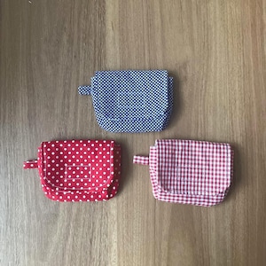 Puede incluir: Tres pequeñas bolsas de tela. Una es roja con lunares blancos, otra es azul con lunares blancos y la tercera es a cuadros rojos y blancos. Cada una tiene una pequeña pestaña en el lateral. Sobre una superficie de madera.