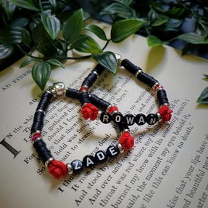 Op de afbeelding: Twee armbanden met de namen "ZADE" en "ROWAN" gespeld in zwarte en witte letterkralen. De armbanden hebben zwarte schijfkralen, rode rozenaccenten en zilverkleurige kralen. De armbanden worden tentoongesteld op een open boek.