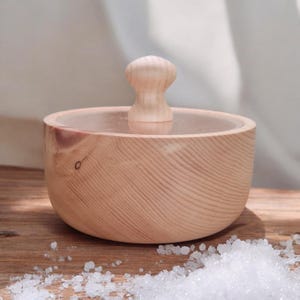 Könnte beinhalten: Ein hölzerner Salzstreuer mit Deckel und kleinem Holzgriff. Der Salzstreuer ist aus hellem Holz gefertigt und hat ein glattes, geschwungenes Design. Grobe Salzkristalle sind auf der Holzoberfläche verstreut.