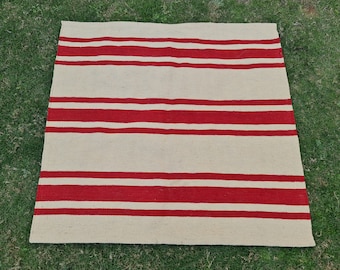 Puede incluir: Una alfombra cuadrada que presenta un patrón repetitivo de rayas horizontales rojas sobre un fondo beige natural. El tejido tiene una superficie plana con bordes rectos y limpios, y una distribución de color consistente en toda la pieza.