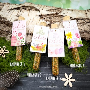 May include: Four glass tubes filled with spices, each capped with a cork and a decorative tag. The tags feature Easter-themed designs with text in German. The tubes are arranged on a bed of green moss, with wooden accents.