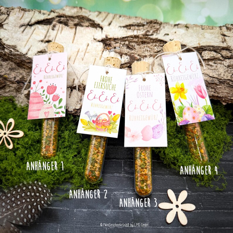 May include: Four glass tubes filled with spices, each capped with a cork and a decorative tag. The tags feature Easter-themed designs with text in German. The tubes are arranged on a bed of green moss, with wooden accents.