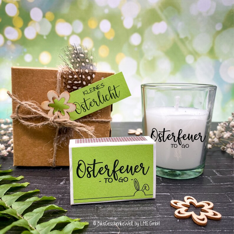 May include: A green and white box with the text "Osterfeuer - TO GO" and a small bunny rabbit graphic. A clear glass candle holder with a white candle inside. The candle holder has the text "Osterfeuer TO GO" on the side. The items are on a wooden surface with green foliage.