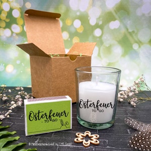 May include: A small glass candle jar with a white candle and the text "Osterfeuer to go" on the side. The candle is next to a matchbox with the same text and a small wooden flower.