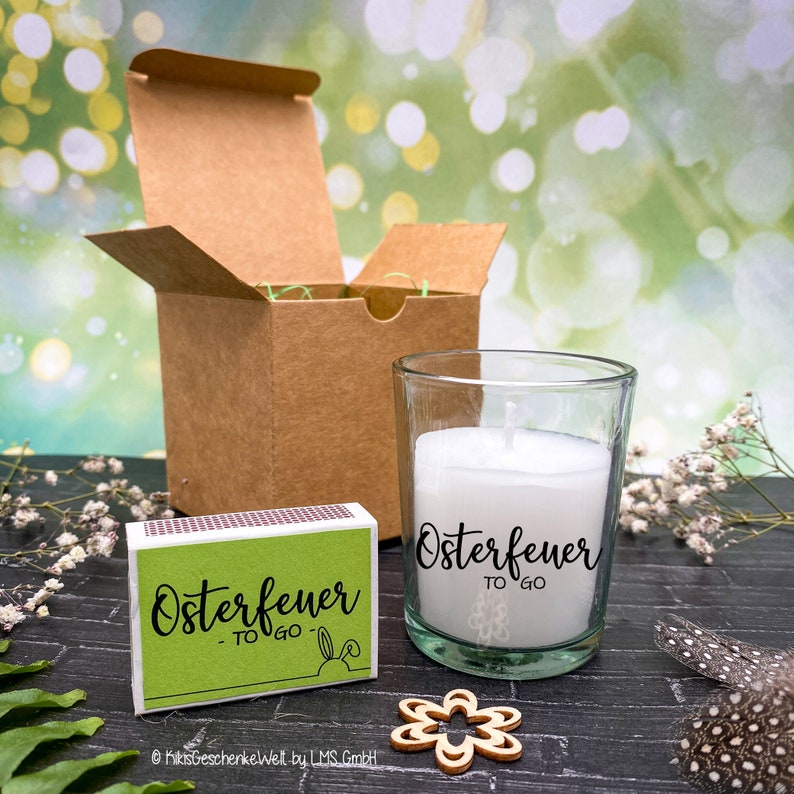 May include: A small glass candle jar with a white candle and the text "Osterfeuer to go" on the side. The candle is next to a matchbox with the same text and a small wooden flower.