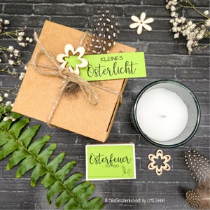 May include: A brown gift box with a green tag that says "Kleines Osterlicht" and a wooden flower decoration. The box is tied with twine and a feather is on top. A small white candle in a glass jar and a green matchbox with the text "Osterfeuer - to go -" are also in the image.