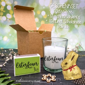May include: A gift set with a brown box, a glass candle with a white candle and a green label that says "Osterfeuer TO GO", a box of matches with a green label that says "Osterfeuer TO GO", and a gold foil wrapped chocolate bunny with a red ribbon.