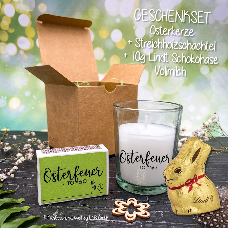 May include: A gift set with a brown box, a glass candle with a white candle and a green label that says "Osterfeuer TO GO", a box of matches with a green label that says "Osterfeuer TO GO", and a gold foil wrapped chocolate bunny with a red ribbon.