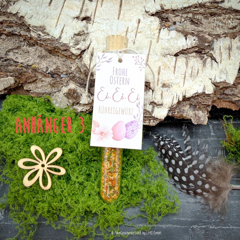May include: A glass vial filled with spices, topped with a cork, and a tag that reads "Frohe Ostern" with egg graphics. The vial is set on green moss with a wooden flower and a speckled feather.