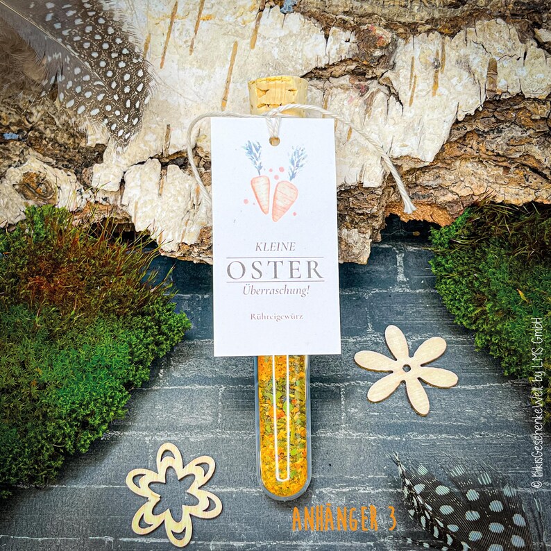 May include: A glass vial filled with yellow and green granules, topped with a cork and a tag that reads "Kleine Oster &Uuml;berraschung!" with carrot illustrations. The vial is suspended against a backdrop of wood, moss, and decorative wooden flowers.