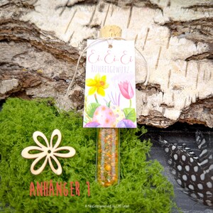 May include: A clear glass vial filled with spices, topped with a cork and a tag. The tag features the text "Ei. Ei. Ei. RUHRGEWURZ" and a watercolor illustration of Easter eggs and flowers. The vial is set on green moss.