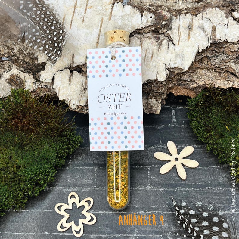 May include: A clear glass vial filled with yellow spices, sealed with a cork, and a tag that reads "OSTER ZEIT". The tag has a polka dot pattern in pink and blue. The vial is displayed with wooden flowers, moss, and feathers.