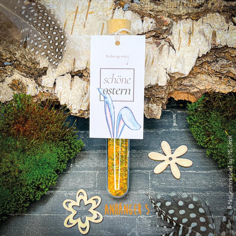 May include: A clear glass vial filled with yellow spices, topped with a cork, and a white tag that reads "sch&ouml;ne Ostern" with bunny ears. The image includes wooden cutouts, feathers, and moss.
