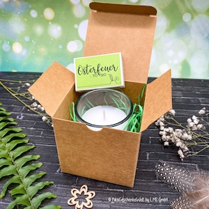 May include: A brown cardboard box with a lid open, revealing a white candle in a clear glass jar. The candle is surrounded by green shredded paper. A green and white sticker with the text "Osterfeuer - TO GO" is on top of the box.