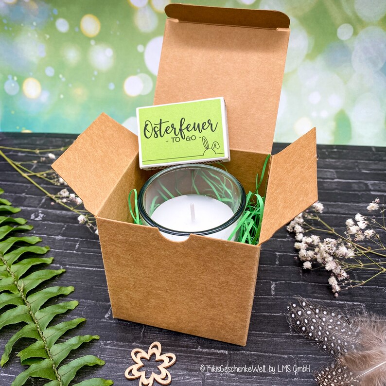 May include: A brown cardboard box with a lid open, revealing a white candle in a clear glass jar. The candle is surrounded by green shredded paper. A green and white sticker with the text "Osterfeuer - TO GO" is on top of the box.