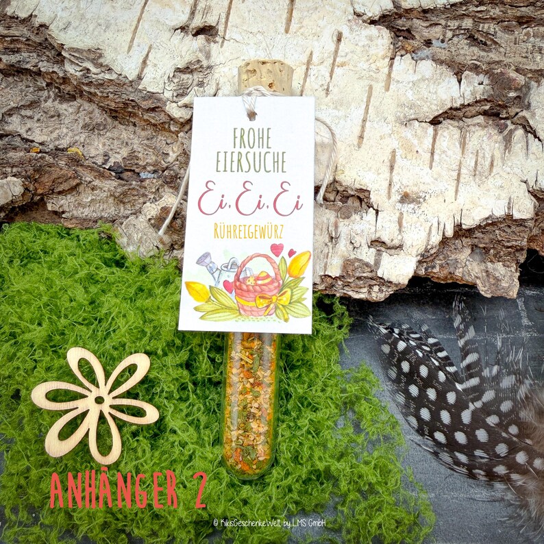May include: A glass vial filled with spices, sealed with a cork, and a tag that reads "Frohe Eiersuche" with a basket illustration. The vial is set on green moss with a wooden flower and a feather.