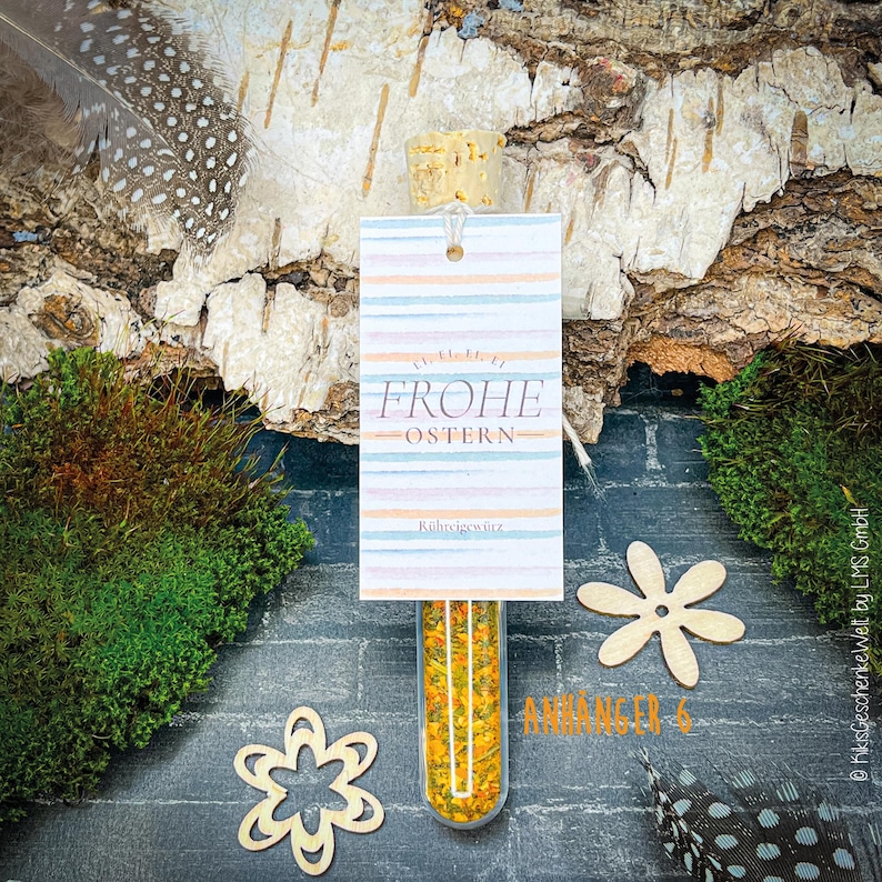 May include: A clear glass tube filled with yellow and orange spices, topped with a cork and a tag that reads "FROHE OSTERN". The tag has pastel stripes. The background includes wood bark, moss, and decorative wooden flowers.