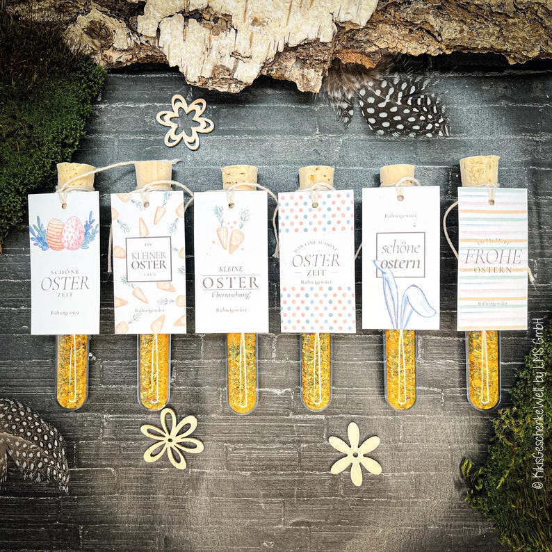 May include: Six glass vials with cork stoppers, each containing yellow granules and a decorative tag. The tags feature Easter-themed designs and text in German. Wooden flowers and feathers are scattered around the vials, creating a festive arrangement.