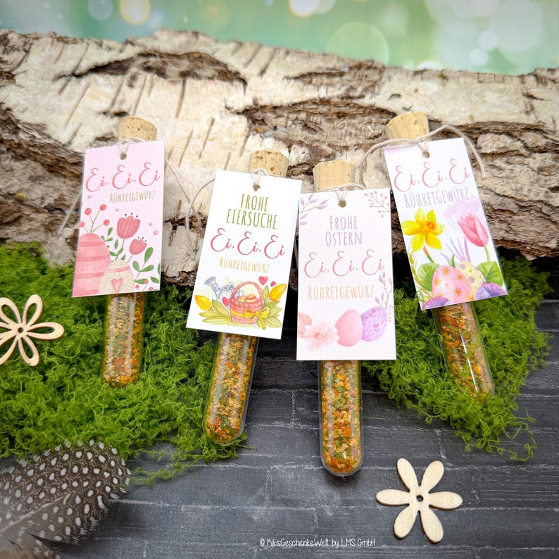 May include: Four glass tubes filled with spices, each capped with a cork and a decorative tag. The tags feature Easter-themed illustrations and German text. The tubes are arranged on a bed of green moss, with a wood background.