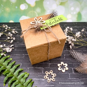 May include: A brown cardboard box with a green tag that says "kleines Osterlicht" is tied with twine and decorated with wooden flower cutouts. The box is sitting on a black brick surface with green ferns and white flowers.