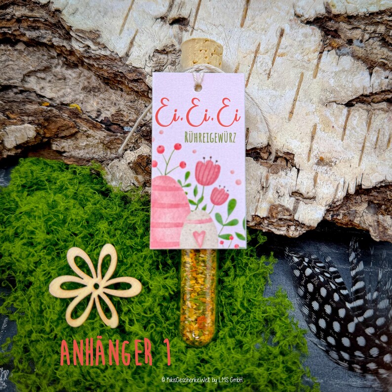 May include: A small glass vial filled with colorful spices, topped with a cork, and a decorative tag. The tag features the text "Ei. Ei. Ei. R&Uuml;HREIGEW&Uuml;RZ" with floral illustrations. The vial is set against a backdrop of green moss and birch bark.