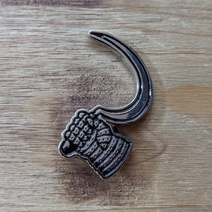 Puede incluir: Un pin de metal plateado con forma de mano sosteniendo un gancho curvo. La mano está detallada con un diseño texturizado.