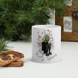 Puede incluir: Taza de cerámica blanca con un personaje de dibujos animados verde con una camisa negra y zapatos marrones. El personaje está rodeado de cucharas marrones. El texto en la taza dice "Me gustan las cucharas oxidadas".