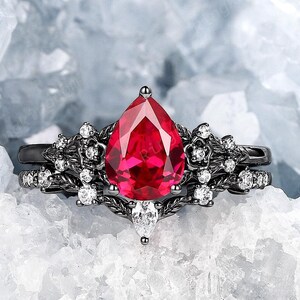 May include: A black ring featuring a pear-shaped red gemstone surrounded by smaller white gemstones. The ring is displayed against a white backdrop.