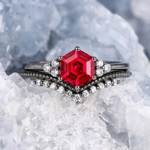 May include: A black ring set with a large, red, hexagonal gemstone surrounded by smaller, clear gemstones. The ring is set on a white, crystalline background.