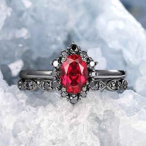 Puede incluir: Un conjunto de anillos negros con una gran piedra roja ovalada rodeada de piedras negras más pequeñas. El anillo está sobre un fondo blanco.