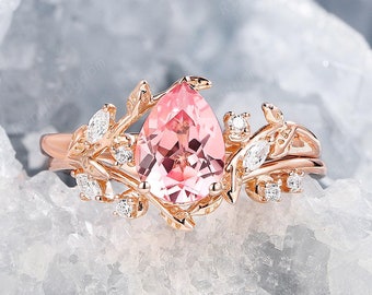 Könnte beinhalten: Ein Verlobungsring aus Roségold mit einem birnenförmigen rosafarbenen Edelstein, der von kleineren Diamanten umgeben ist. Der Ring hat ein zartes florales Design mit Blättern und Ranken.