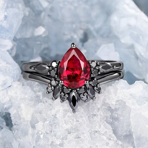 May include: A black ring featuring a pear-shaped red gemstone surrounded by smaller black gemstones. The ring is displayed against a white backdrop.